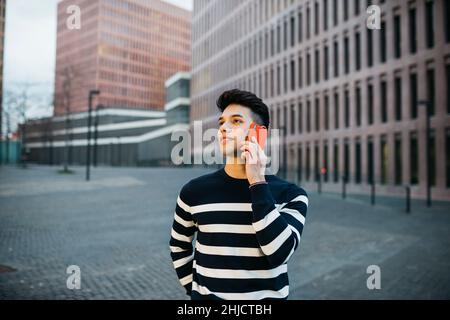 Junger lächelnder Mann, der auf einem roten Smartphone in der A-Straße läuft und spricht Stockfoto