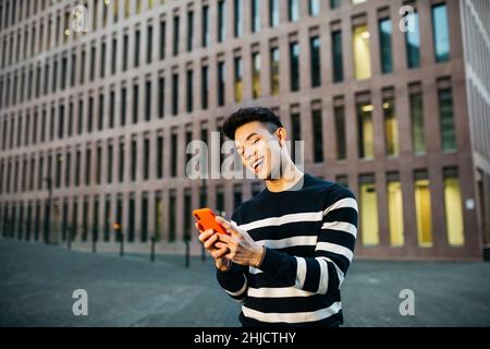 Junger lächelnder Mann, der auf der Straße mit einem roten Smartphone unterwegs ist Stockfoto