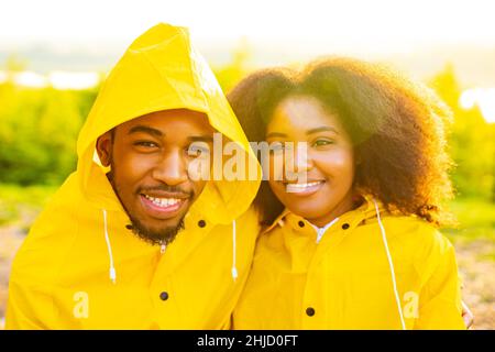 afroamerikanische Frau und Ehemann in gelbem Regenmantel mit Kapuze, die sich bei Sonnenuntergang im Freien glücklich fühlen Stockfoto