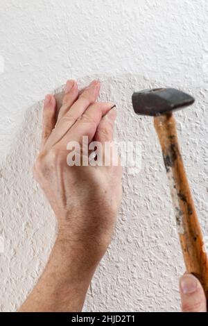 Nahaufnahme einer Hand mit einem Stahlnagel, der den Nagel mit einem Hammer in eine tapeerte weiße Wand treibt. Stockfoto