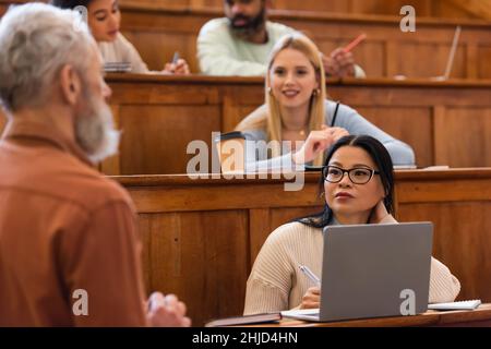 Asiatische Student Blick auf verschwommene Lehrer in der Nähe von Laptop und Notebooks in der Universität Stockfoto