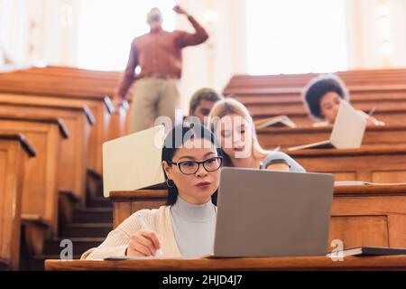 Asiatische Studenten schreiben auf Notebook in der Nähe von Laptop und verschwommen multiethnischen Freunden in der Universität Stockfoto