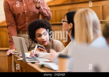 afroamerikanische Studentin, die verschwommene Freunde in der Nähe von Laptops und Lehrern in der Universität ansieht Stockfoto