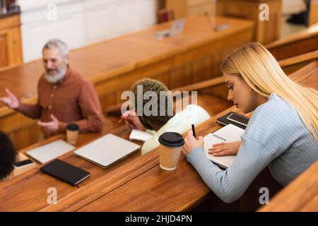Student schrieb auf Notebook in der Nähe von Kaffee zu gehen und verschwommen interracial Freunde in der Universität Stockfoto