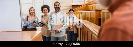 Positive interracial Studenten mit Notebooks und Geräten Blick auf verschwommene Lehrer in der Universität, Banner Stockfoto