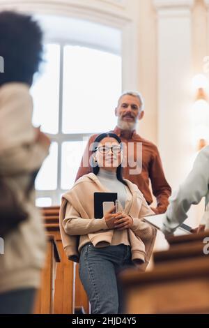 Low-Angle-Ansicht eines lächelnden asiatischen Studenten mit Smartphone und Notebook, der die Kamera in der Nähe von verschwommenen Freunden und Lehrern in der Universität ansieht Stockfoto