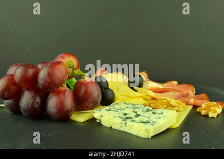 Verschiedene Käsesorten auf schwarzem Schieferbrett. Brie-Käse, Cheddar-Scheiben, Walnüsse, Trauben, Oliven und Schwarzwald-Schinken. Stillleben Essen. Speicherplatz kopieren Stockfoto