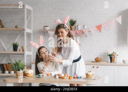 Eine schöne junge Mutter hilft ihrer kleinen Tochter, Ostereier in der heimischen Küche zu malen. Festliche Stimmung. Stockfoto