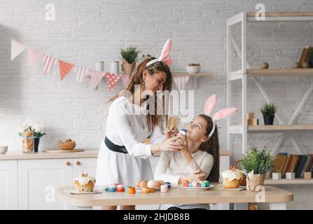 Eine schöne junge Mutter hilft ihrer kleinen Tochter, Ostereier in der heimischen Küche zu malen. Festliche Stimmung. Stockfoto