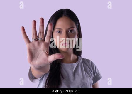 Junge latinerin im lässigen Outfit, die das Hand-Stop-Zeichen vor fliederem Studiohintergrund macht.Nahaufnahme und selektiver Fokus, Kopierraum. Missbrauch stoppen, Disc Stockfoto