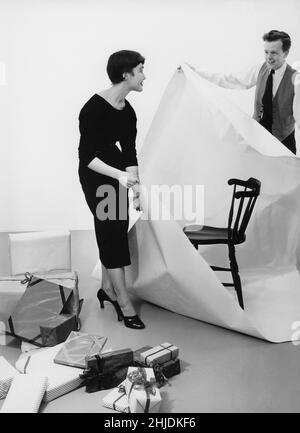 Weihnachten im Jahr 1950s. Ein Paar mit sichtbaren Weihnachtsgeschenken versucht sein Bestes, einen Stuhl in Papier zu wickeln. 1953 Stockfoto
