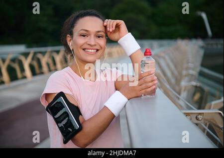 Attraktive junge Sportlerin mit Smartphone-Halterung am Arm, lächelt mit einem schönen Lächeln und schaut auf die Kamera, während sie sich auf der Stadtbrücke ausruht Stockfoto