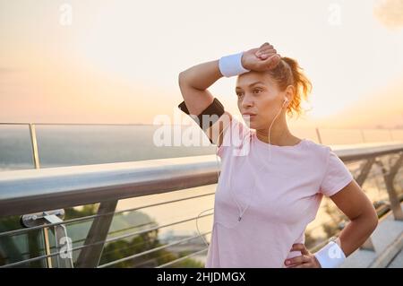 Eine müde Athletin, erschöpft Sportlerin wischt Schweiß von ihrer Stirn mit terry weißen Armbändern, steht auf der Stadtbrücke, nach einem ausruhen Stockfoto