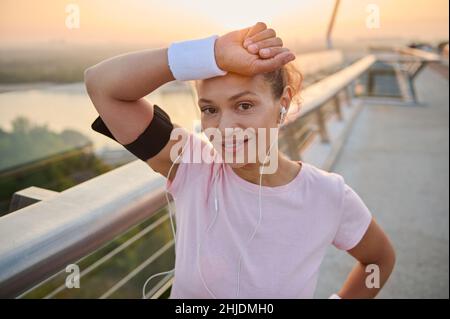 Die schöne hispanische Sportlerin, erschöpfte Sportlerin, wischt sich mit Frottee-Armbändern Schweiß von der Stirn, lächelt und blickt auf die Kamera, die auf der steht Stockfoto