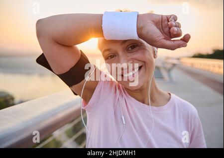 Headshot einer schönen hispanischen Athletin, die mit einem Frottee-Armband Schweiß von der Stirn abwischt, lächelt mit einem fröhlichen, toothy Lächeln, das nach m ausruht Stockfoto