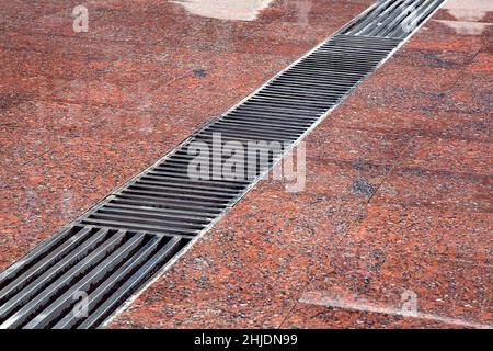 Drainage Rost mit Streifen auf dem Boden mit einer feuchten roten Granitoberfläche, Nahaufnahme eines nach Regen nassen Sturmsystems. Stockfoto