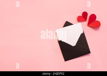 Schwarzer Papierumschlag mit weißem Notizmockup innen und Valentines Hearts auf rosa Hintergrund. Stockfoto