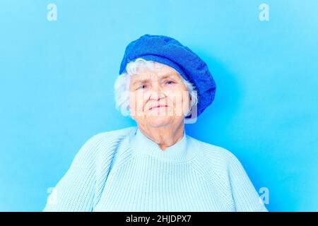 Schöne pensionierte Frau in blauer Baskenmütze und warmer Pullover Blick auf die Kamera und lächeln in blauen Wand Hintergrund im Studio Stockfoto