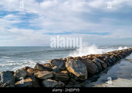 Felsbarrieren Reihen sich entlang des Strandsands zu einer Meereswand mit großen Felsen, die hoch mit blauem bewölktem Himmel im Hintergrund gestapelt sind. Stockfoto