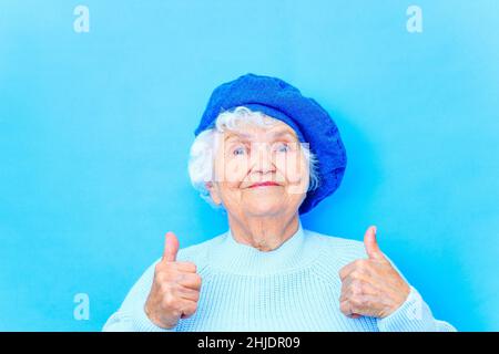 Schöne pensionierte Frau in blauer Baskenmütze und warmer Pullover Blick auf die Kamera und lächeln in blauen Wand Hintergrund im Studio Stockfoto