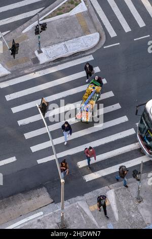 Lateinamerika. Erhöhte Ansicht von Fußgängern auf einem Kreuzweg in Brasilien. Anbieter schiebt einen Obstkorb. Zebra überquert auf der belebten städtischen Avenue. Stockfoto