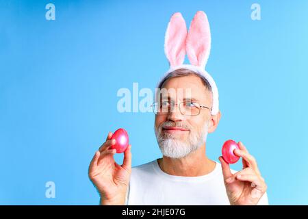 Niedlicher reifer Mann mit Hasenohren, die Osterei auf blauem Hintergrund halten Stockfoto