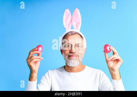 Niedlicher reifer Mann mit Hasenohren, die Osterei auf blauem Hintergrund halten Stockfoto