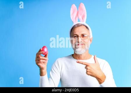 Niedlicher reifer Mann mit Hasenohren, die Osterei auf blauem Hintergrund halten Stockfoto