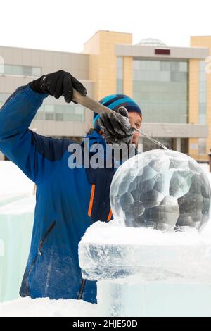 Ein Bildhauer schnitzt mit einem Meißel eine runde Eiskugel aus einem Eisblock. Stockfoto