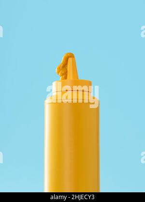 Nahaufnahme mit gelber Senfflasche vor blauem Hintergrund. Ein Tropfen Senf oben auf der Flasche. Gelbe Kunststoffflasche isoliert auf b Stockfoto