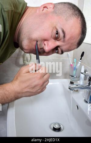 Ein weißer Mann putzt sich die Zähne in der Badewanne über dem Waschbecken. Er schaut in die Kamera. Der Wasserhahn ist offen und Wasser fließt. Stockfoto