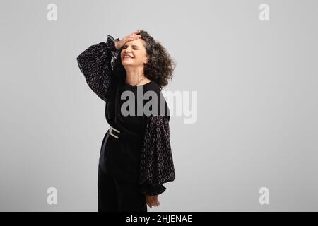Sehr glückliche ältere Frau mit üppigem lockigem Haar, die im Studio auf grauem Hintergrund lachte Stockfoto