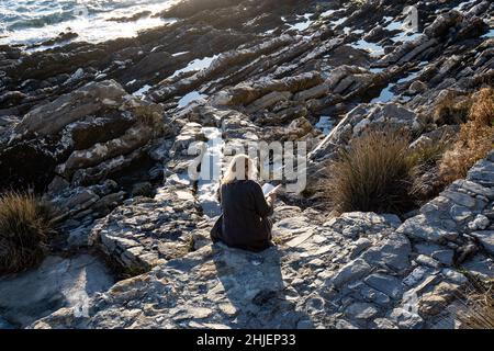 Italien, Genua - Eine junge Frau, die auf den Klippen sitzt, liest ein Buch und entspannt sich im warmen Licht des Sonnenuntergangs im alten Dorf Nervi Stockfoto