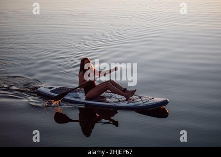 Glückselige Frau mittleren Alters sitzt auf dem Brett Rudern mit Ruder auf dem See mit klarem blauen Wasser lächelt mit Zähnen und entspannend. Von oben aufgenommen. Aktiv Stockfoto