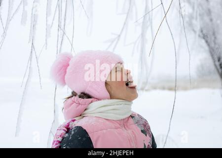 Kaukasische glückliche junge Mädchen versuchen, Schneeflocken mit Zunge mit geschlossenen Augen mit Baumzweigen und schneebedeckten Feld im Hintergrund zu fangen. Winterspaziergang im Park Stockfoto