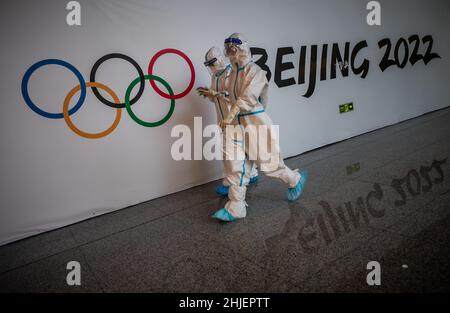 Peking, China. 29th Januar 2022. Helfer in voller Schutzausrüstung laufen an den Olympischen Ringen und dem Schriftzug „Beijing 2022“ vorbei. Athleten, Delegationen und Journalisten, die zu den Olympischen Winterspielen 2022 mit speziellen Charterflugzeugen anreisen, werden mit speziellen Sicherheitsmaßnahmen behandelt. Quelle: Michael Kappeler/dpa/Alamy Live News Stockfoto