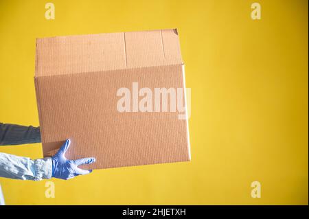 Nahaufnahme der weiblichen Hände in Handschuhen und einem Jeanshemd. Der Lieferer übergibt dem Kunden auf gelbem Hintergrund einen Karton. Antimikrobiell Stockfoto