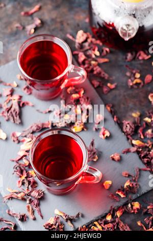 Hibiskus-Tee in Glaskrug auf dunklem Hintergrund. Tasse roten Hibiskustee und trockene Hibiskusblüten auf einer Schiefertafel Stockfoto