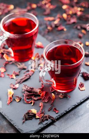 Hibiskus-Tee in Glaskrug auf dunklem Hintergrund. Tasse roten Hibiskustee und trockene Hibiskusblüten auf einer Schiefertafel Stockfoto