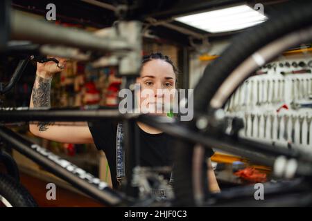 Konzentrierte junge tätowierte weibliche Master mit dunklen Haaren Fixierung modernen Fahrrad während der Arbeit in Reparatur-Garage Stockfoto