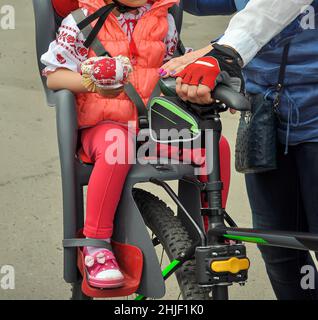 Kinderfahrradsitz aus grauem Kunststoff. Ein Kind ohne Gesicht in roter Kleidung sitzt auf einem Fahrrad, die Hände der Eltern sind sichtbar Stockfoto