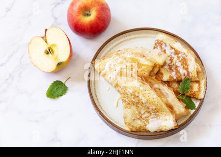 Wir feiern den Pfannkuchen-Tag, ein gesundes Frühstück. Hausgemachte süße Crepes mit Äpfeln und Kondensmilch auf einer Marmorplatte. Stockfoto