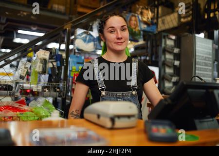 Freundliche junge tätowierte Verkäuferin in legerer Kleidung mit grünen Dreadlocks, die lächeln und die Kamera betrachten, während sie an der Holztheke mit Bargeld steht Stockfoto
