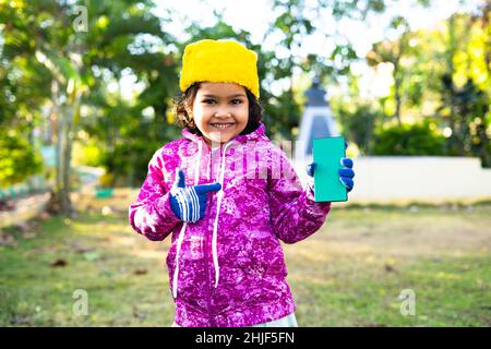 Lächelndes Mädchen Kind zeigt Handy mit grünem Bildschirm Mockup durch Blick auf camea - Konzept der App-Werbung, Winter-Aktionen und Technologie. Stockfoto