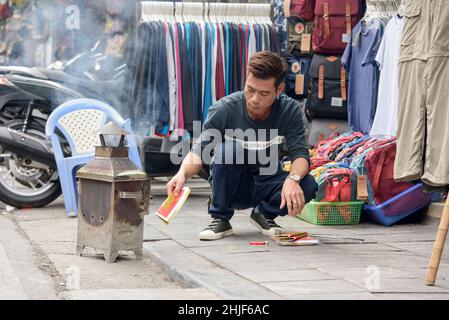 Ein Vietnamese verbrennt Votivgeld (Scherzpapier, gefälschtes Geld, Geistergeld) in einem Brenner auf den Straßen der Altstadt von Hanoi, Vietnam Stockfoto