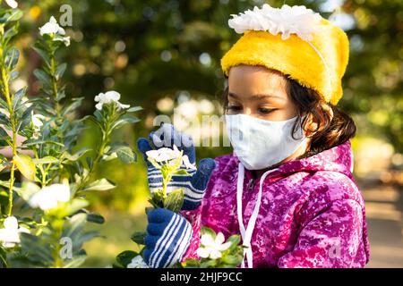 Mädchen Kind mit medizinischer Gesichtsmaske spielt mit Blumen im Park - Konzept der Gesundheitsversorgung, Umweltverschmutzung Sicherheitsmaßnahmen, Achtsamkeit und covid-19 Schutz. Stockfoto