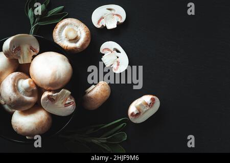 Ganze und schneiden Champignons und Kräuter auf schwarzem Hintergrund. Nahaufnahme, flach liegend, Draufsicht. Lebensmittelhintergrund, selektiver Fokus. Stockfoto