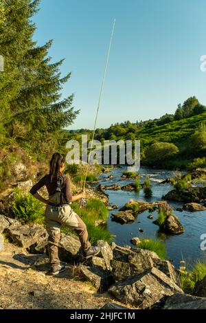 Eine asiatische weibliche Fliegenfischer Frauen, die Watvögel tragen und eine Rute halten und suchen, wo sie auf einem Fluss fischen können Stockfoto