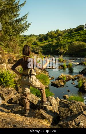 Eine asiatische weibliche Fliegenfischer Frauen, die Watvögel tragen und eine Rute auf ihrer Schulter ruhen, während sie schaut, wo sie auf einem Fluss in Schottland fischen kann Stockfoto