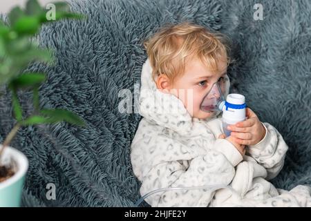 Niedlicher Baby Junge macht Inhalation mit einem Vernebler Ausrüstung. Krankes Kind, das den Inhalator in der Hand hält und zu Hause durch einen Inhalator atmet. Physische Stockfoto